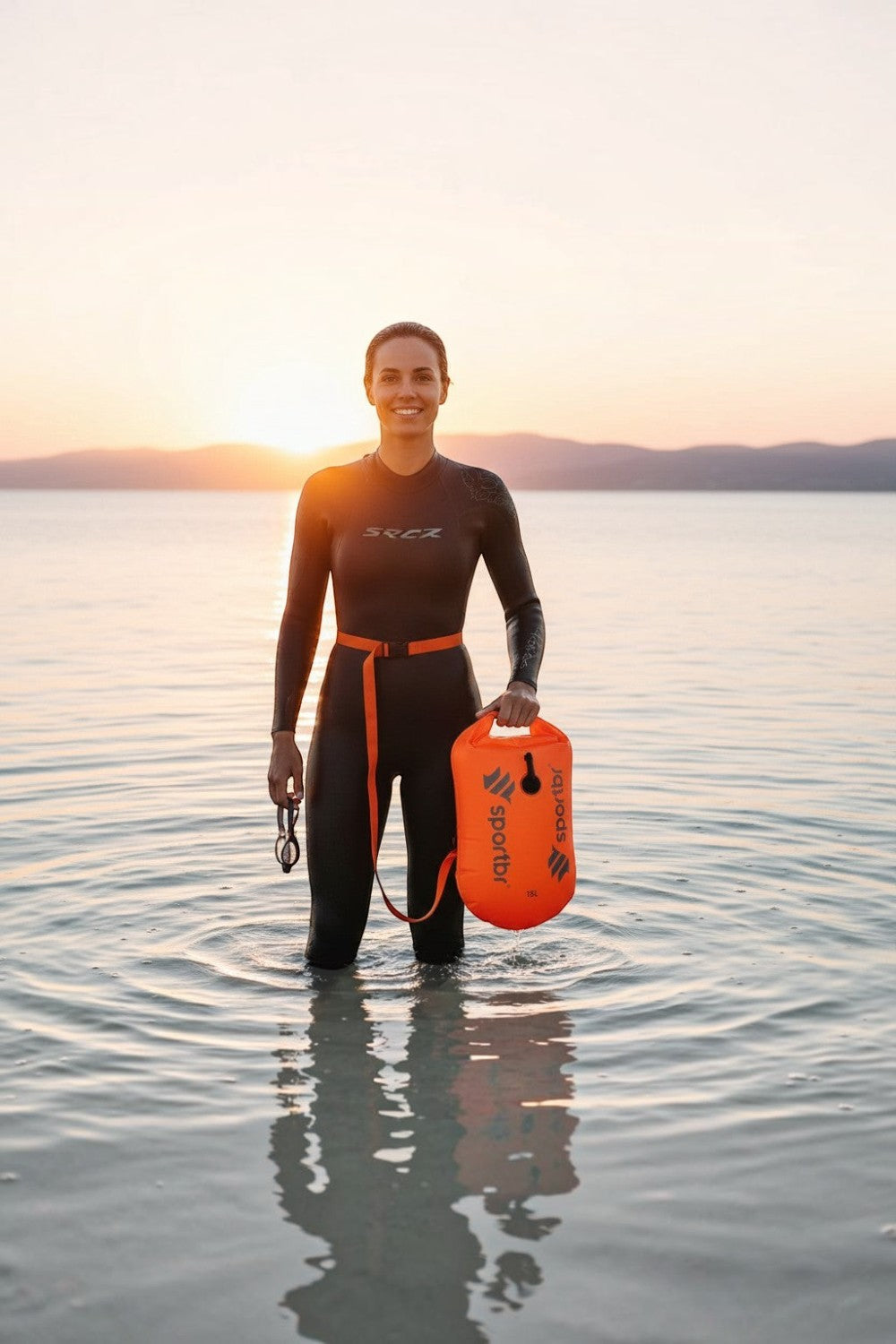 Nadadora com roupa de borracha segurando bóia de sinalização laranja SPORTBR de 15L em águas abertas ao pôr do sol