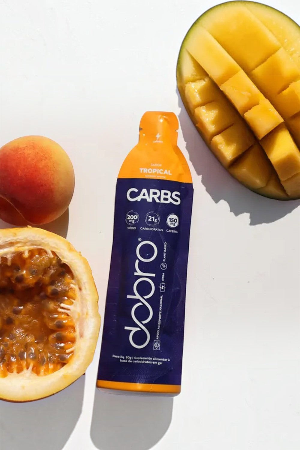 Sachê de gel de carboidrato SPORTBR Dobro sabor tropical com cafeína, ao lado de uma manga e um maracujá frescos.