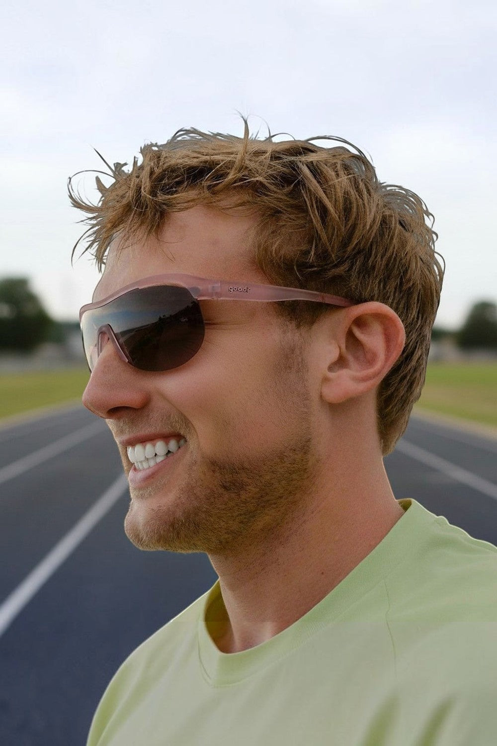 Homem sorrindo usando óculos de corrida SPORTBR Goodr Human Energy Drink com armação rosa translúcida em pista de atletismo