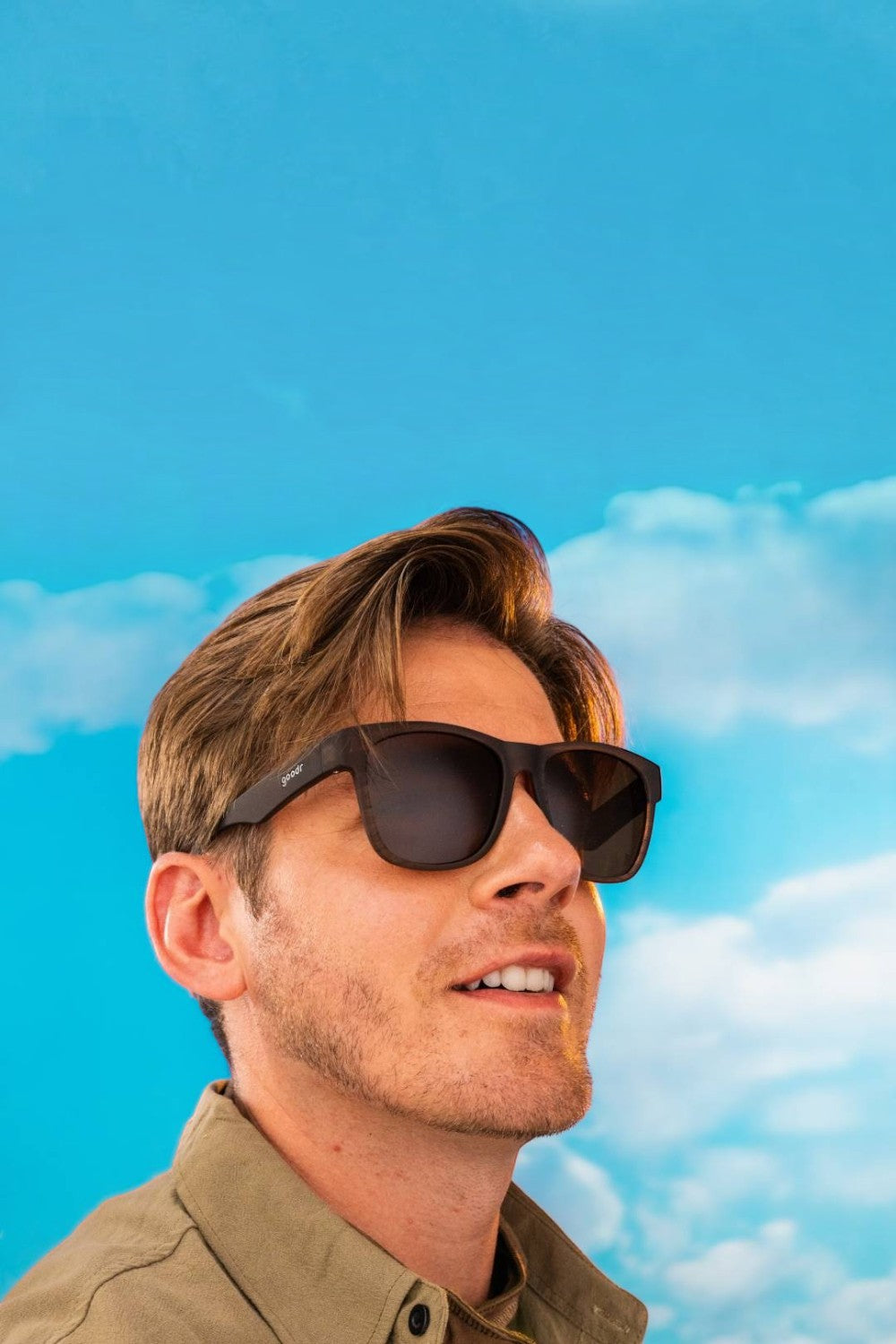 Homem usando óculos de sol SPORTBR Goodr BFG marrons com lentes polarizadas contra um céu azul ensolarado.
