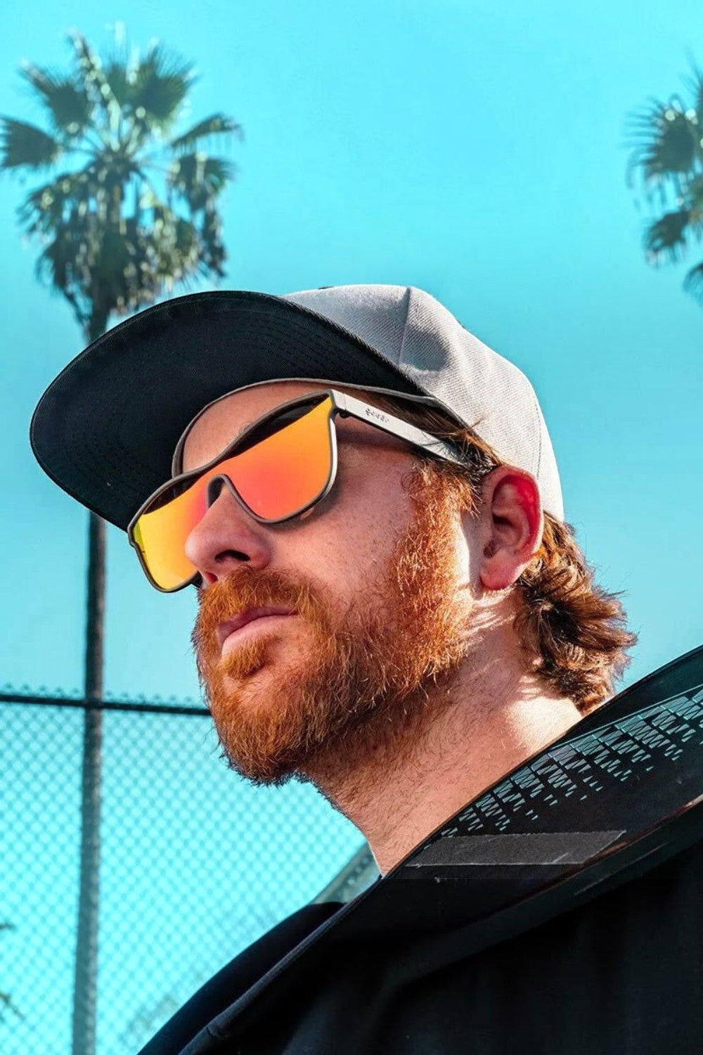 Homem ruivo usando óculos de sol SPORTBR Goodr Voight-Kampff Vision com armação cinza e lentes espelhadas laranja.
