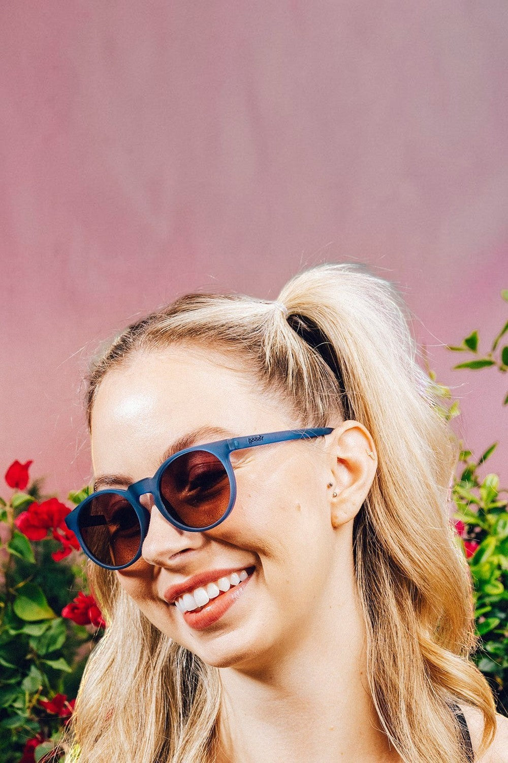 Mulher sorridente usando óculos de sol SPORTBR Goodr Stares Into the Abyss com armação azul e lentes âmbar polarizadas.