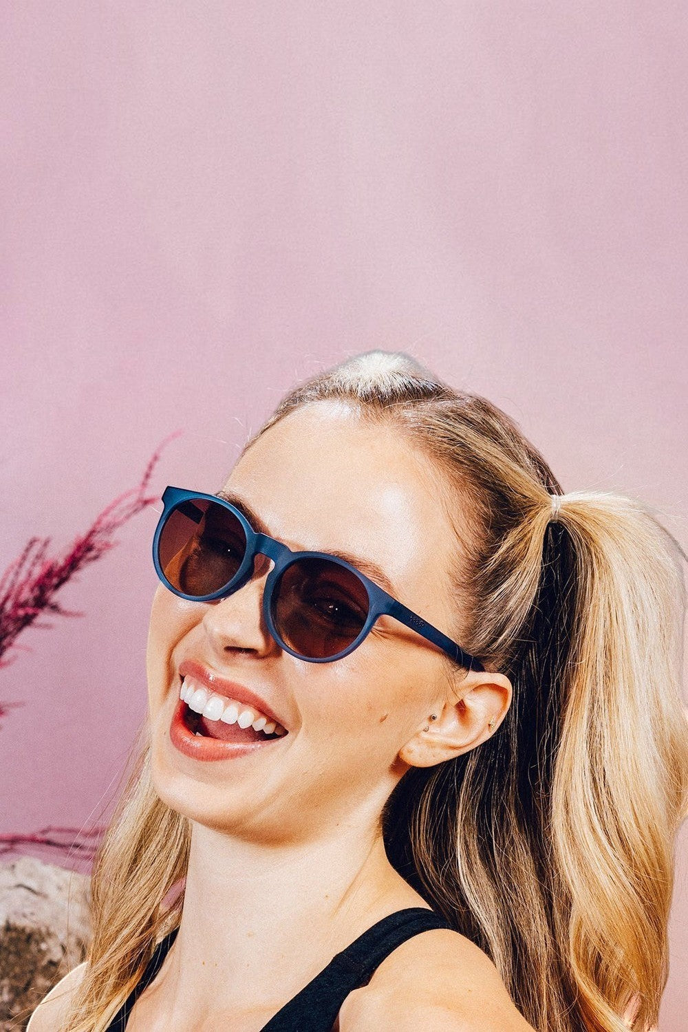 Mulher sorridente usando óculos de sol SPORTBR Goodr Stares Into the Abyss com armação azul e lentes âmbar polarizadas