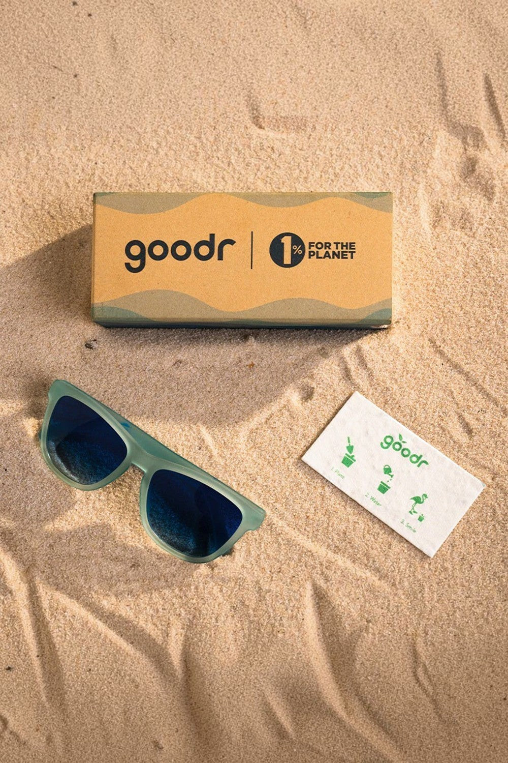 Óculos de sol SPORTBR Goodr azul claro com lentes polarizadas, caixa e cartão informativo sobre areia.