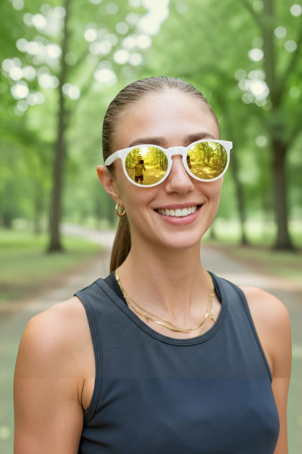 Mulher sorridente usando óculos de sol SPORTBR Goodr Yolk's on You com armação branca e lentes espelhadas amarelas em parque.