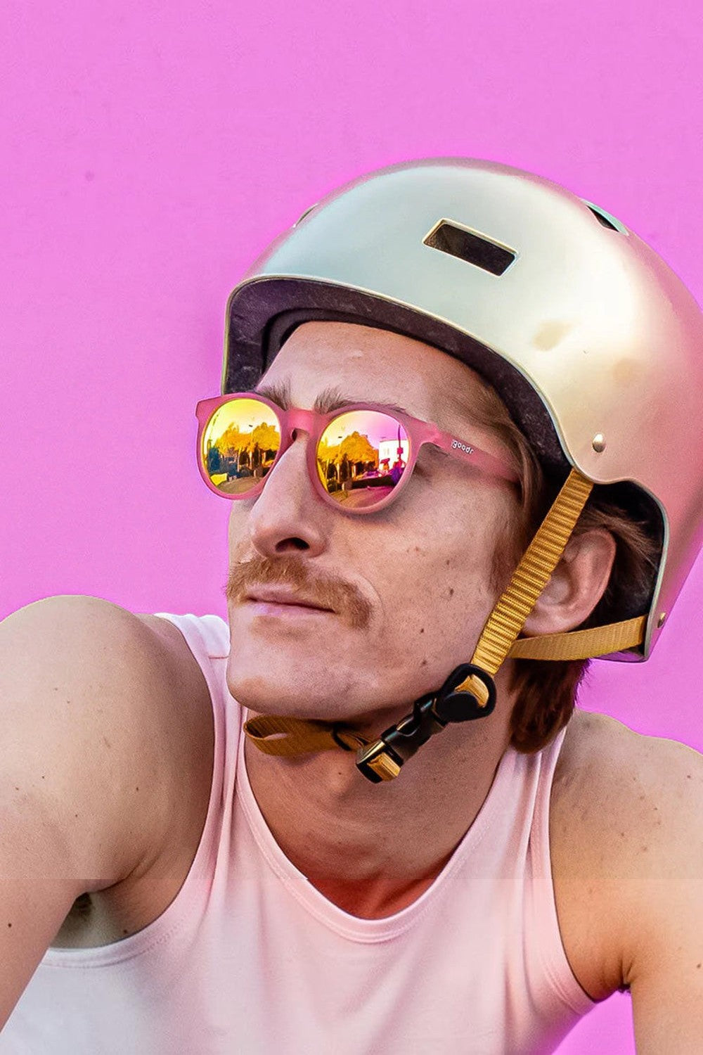 Homem usando óculos de sol SPORTBR Goodr rosa com lentes espelhadas e capacete dourado em fundo rosa.