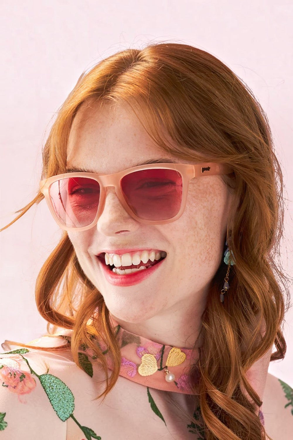 Modelo sorrindo usando óculos de sol SPORTBR Goodr Don't Make Me Blush rosa com lentes polarizadas e armação leve.