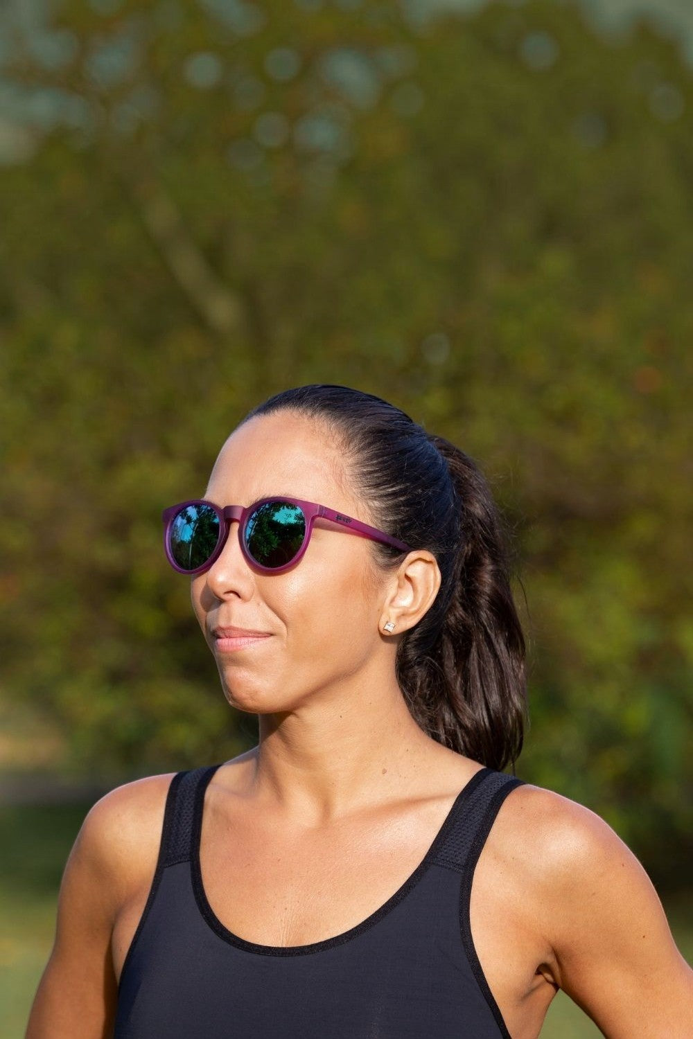 Mulher usando óculos de sol SPORTBR Goodr roxos com lentes verdes espelhadas em ambiente externo de corrida.