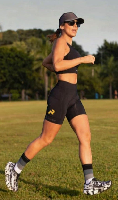 Corredora usando bermuda de compressão preta SPORTBR com bolsos laterais, boné e top esportivo em um campo gramado.