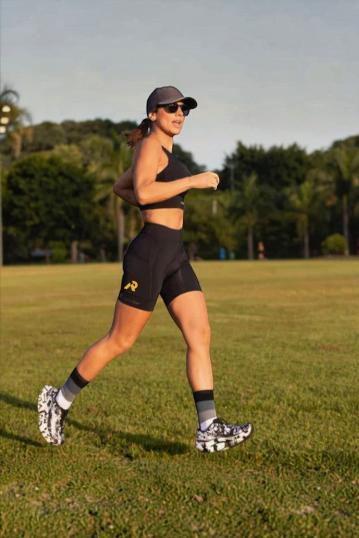 Corredora usando bermuda de compressão preta SPORTBR com bolsos laterais, boné e top esportivo em um campo gramado.