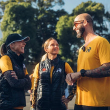 SPORTBR: Três corredores sorriem e conversam ao ar livre, vestindo coletes pretos sobre camisetas amarelas.