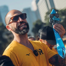 Homem sorridente com óculos de sol e camiseta amarela da SPORTBR, exibindo medalhas de maratona em um dia ensolarado.