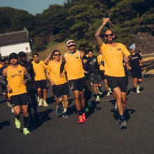 SPORTBR grupo de corredores em uníssono, vestindo camisetas amarelas e shorts pretos, correndo em uma estrada ensolarada.
