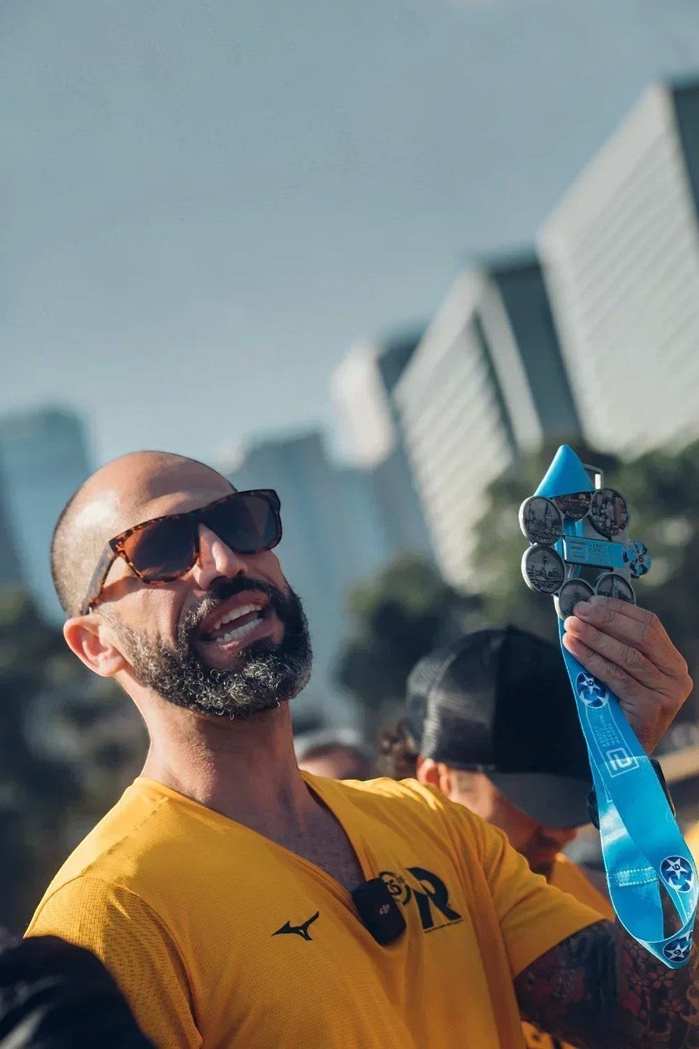 Homem com medalha de maratona e camiseta amarela SPORTBR, celebrando conquista ao ar livre em dia ensolarado.