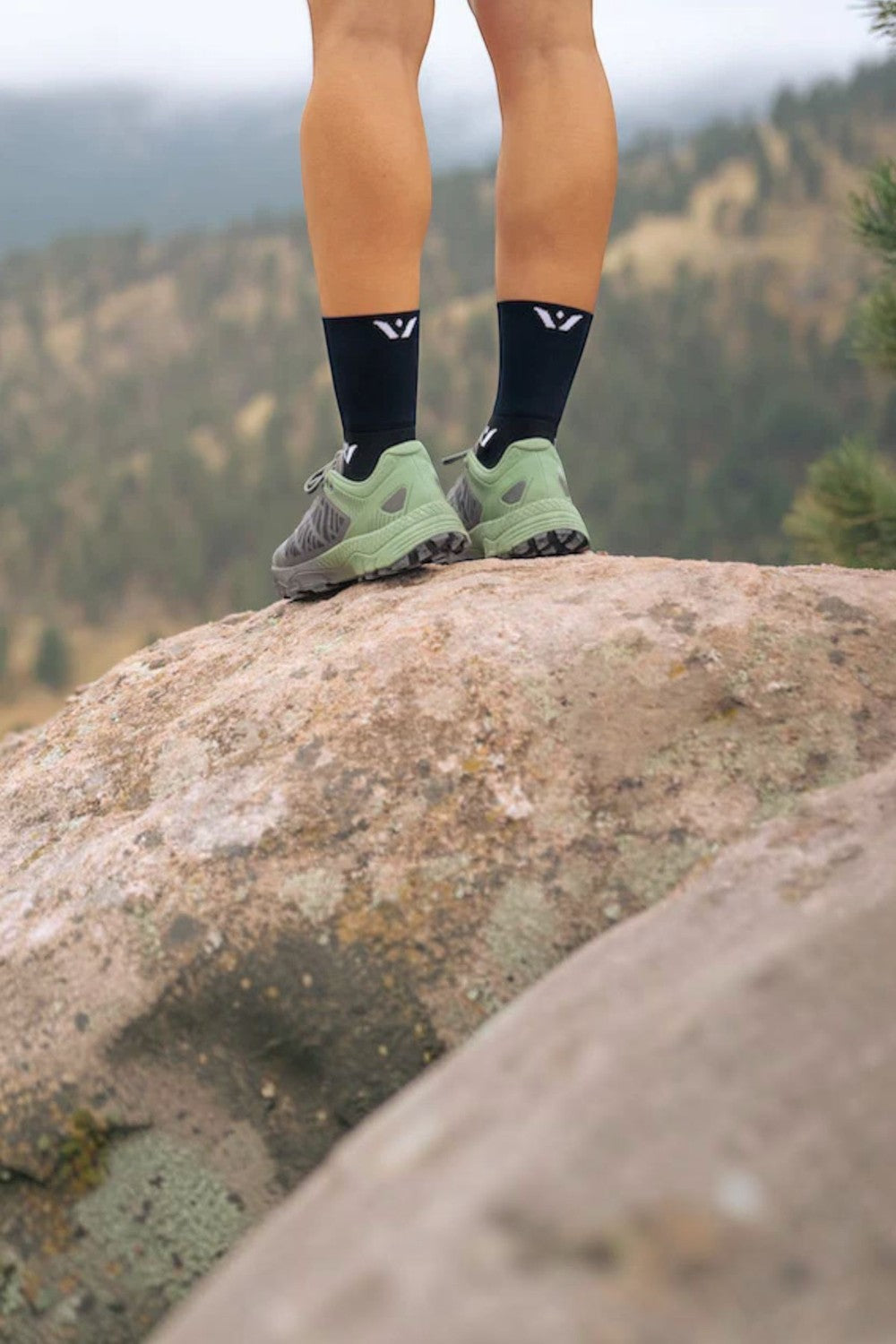 Pernas de corredor usando meia de compressão preta SPORTBR Swiftwick Aspire Four de cano médio e tênis verde em rocha