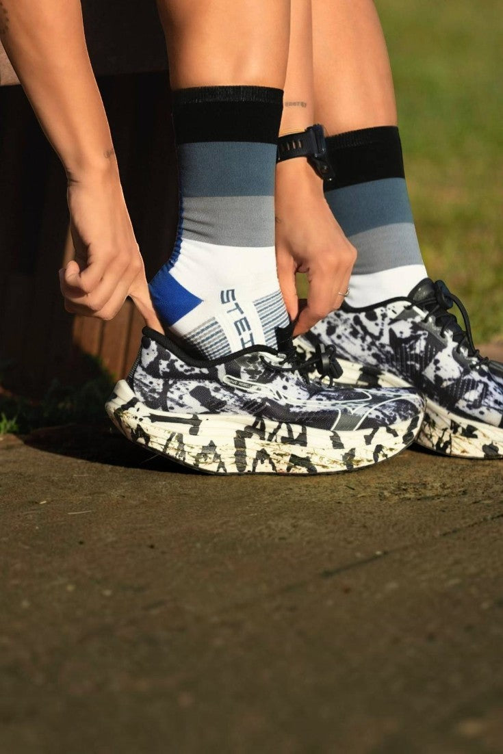 Meia SPORTBR Steigen cano alto Belle com listras cinza, branca e azul, sendo calçada por uma corredora com tênis estampado.
