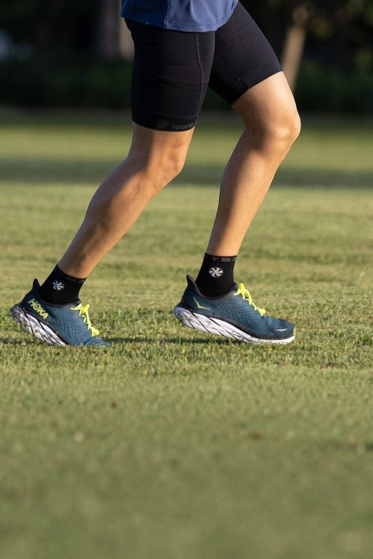 Atleta correndo em gramado usando meia SPORTBR preta de cano médio, tênis Hoka e bermuda esportiva preta.