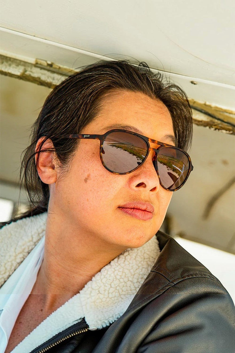 Mulher usando óculos de sol Goodr Amelia Earhart Ghosted Me com armação marrom estilo aviador e lentes polarizadas.