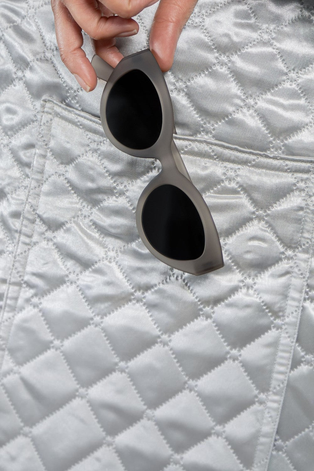 Óculos de sol SPORTBR Goodr Moonbean Daydream cinza com lentes polarizadas, segurados sobre tecido acolchoado prateado.