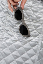 Óculos de sol SPORTBR Goodr Moonbean Daydream cinza com lentes polarizadas, segurados sobre tecido acolchoado prateado.