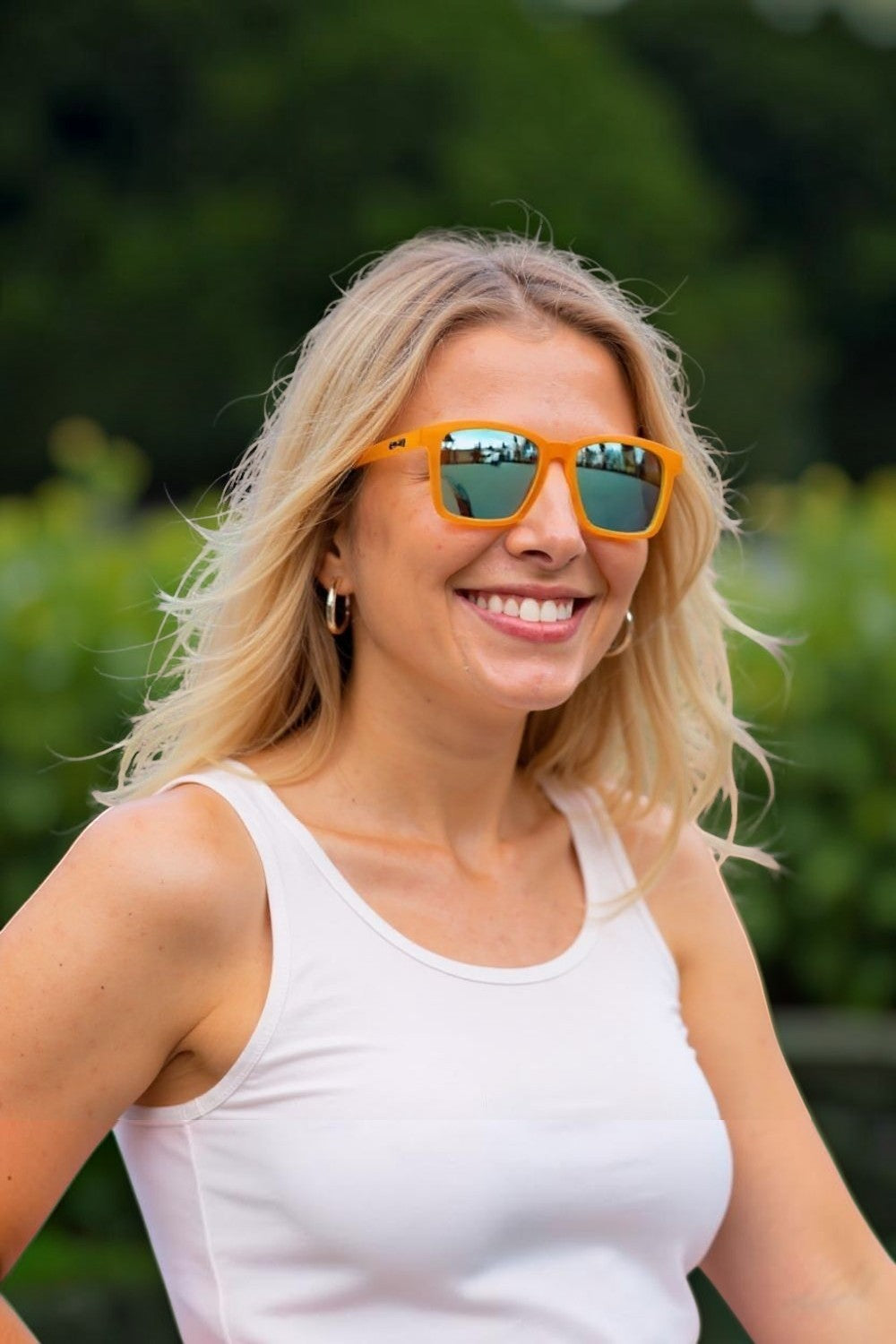 Mulher loira sorrindo usando óculos de sol SPORTBR Goodr laranja com lentes espelhadas azuis em ambiente externo
