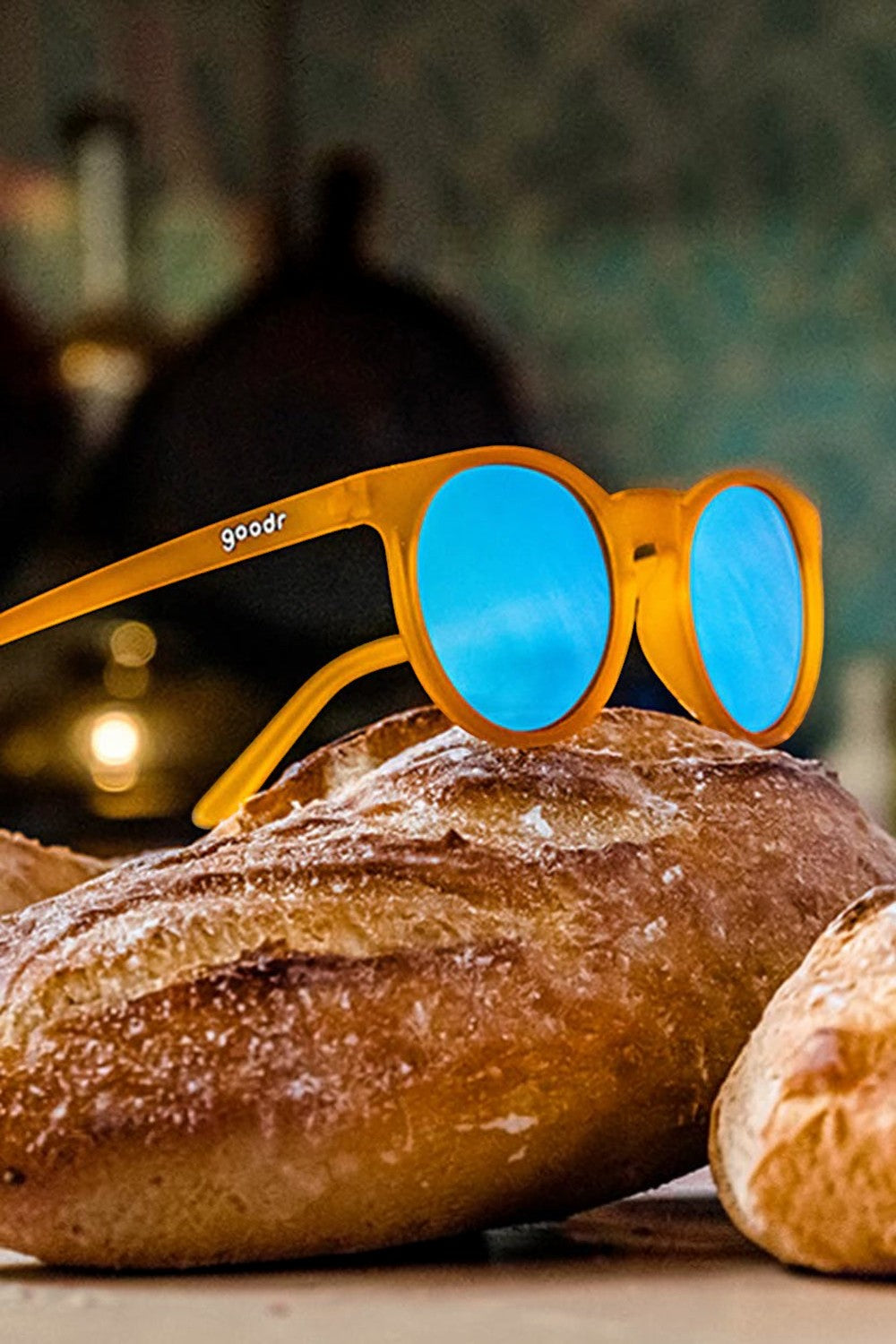 Óculos de sol SPORTBR Goodr laranja com lentes azuis espelhadas, modelo Circle G, apoiados sobre pães artesanais.