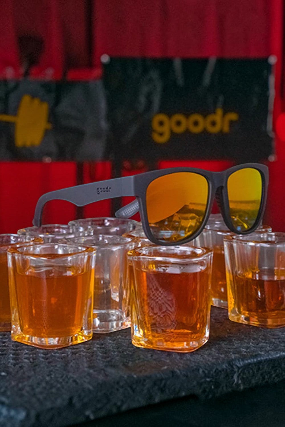Óculos de sol SPORTBR Goodr BFG com armação preta fosca e lentes espelhadas amarelas sobre copos de bourbon.