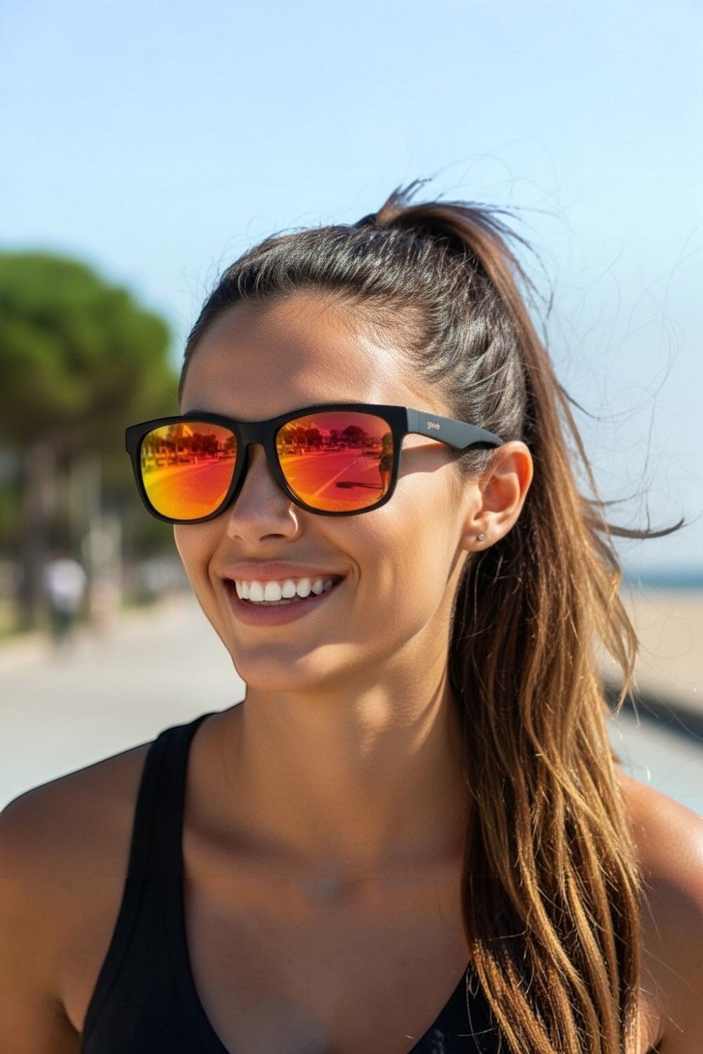Mulher sorridente usando óculos de sol SPORTBR Goodr BFG com armação preta e lentes espelhadas amarelas ao ar livre.