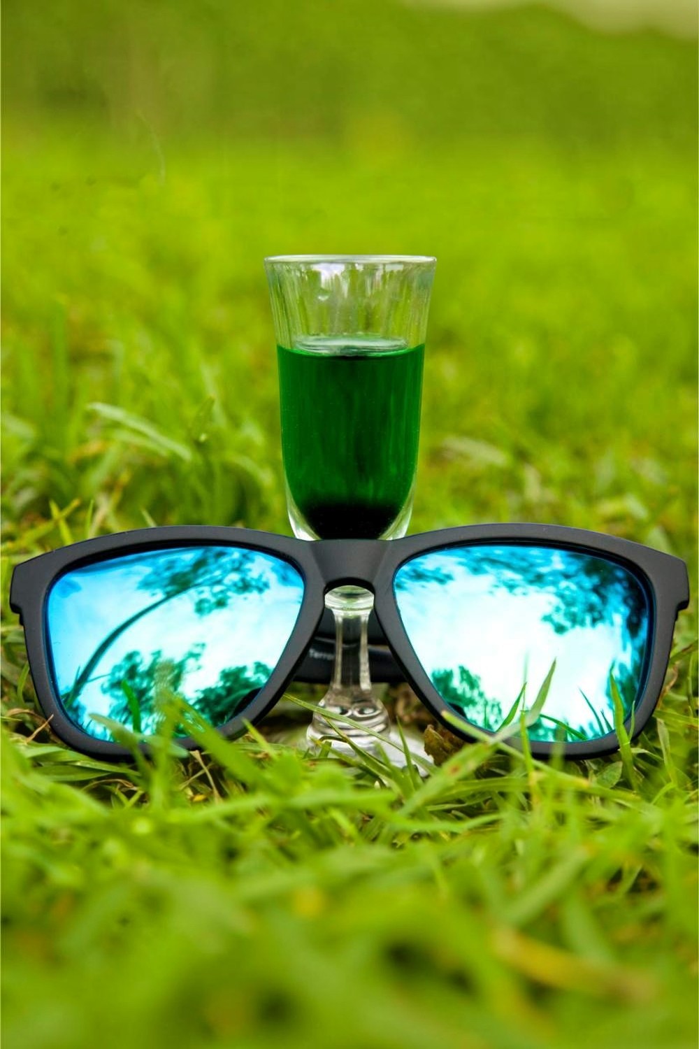 Óculos de sol SPORTBR Goodr pretos com lentes espelhadas verdes sobre gramado, ao lado de uma dose de absinto.