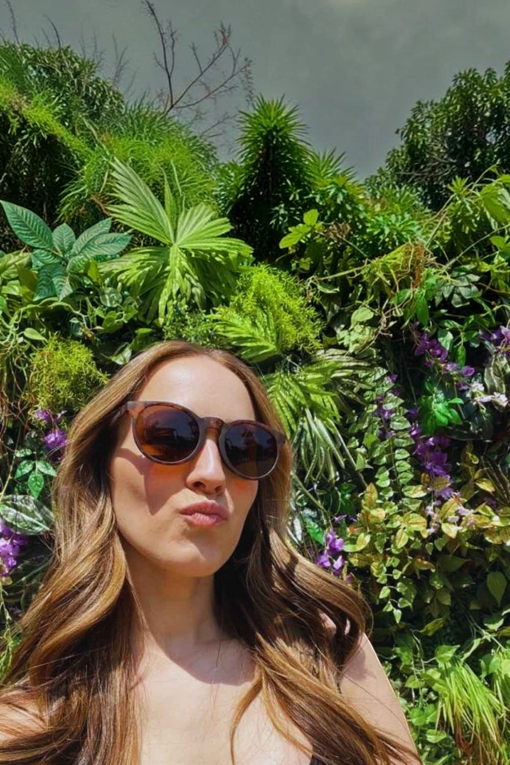 Mulher usando óculos de sol SPORTBR Goodr Nine Dollar Pour Over marrom com lentes polarizadas em um jardim.