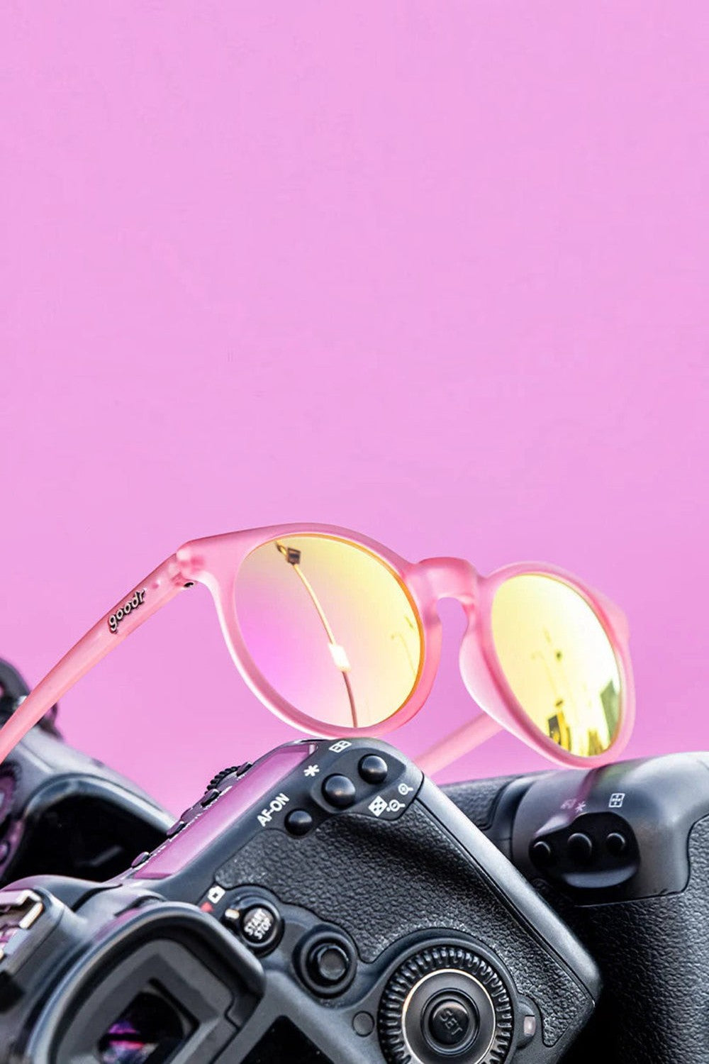 Óculos de sol SPORTBR Goodr Influencers Pay Double rosa com lentes espelhadas polarizadas sobre uma câmera fotográfica.