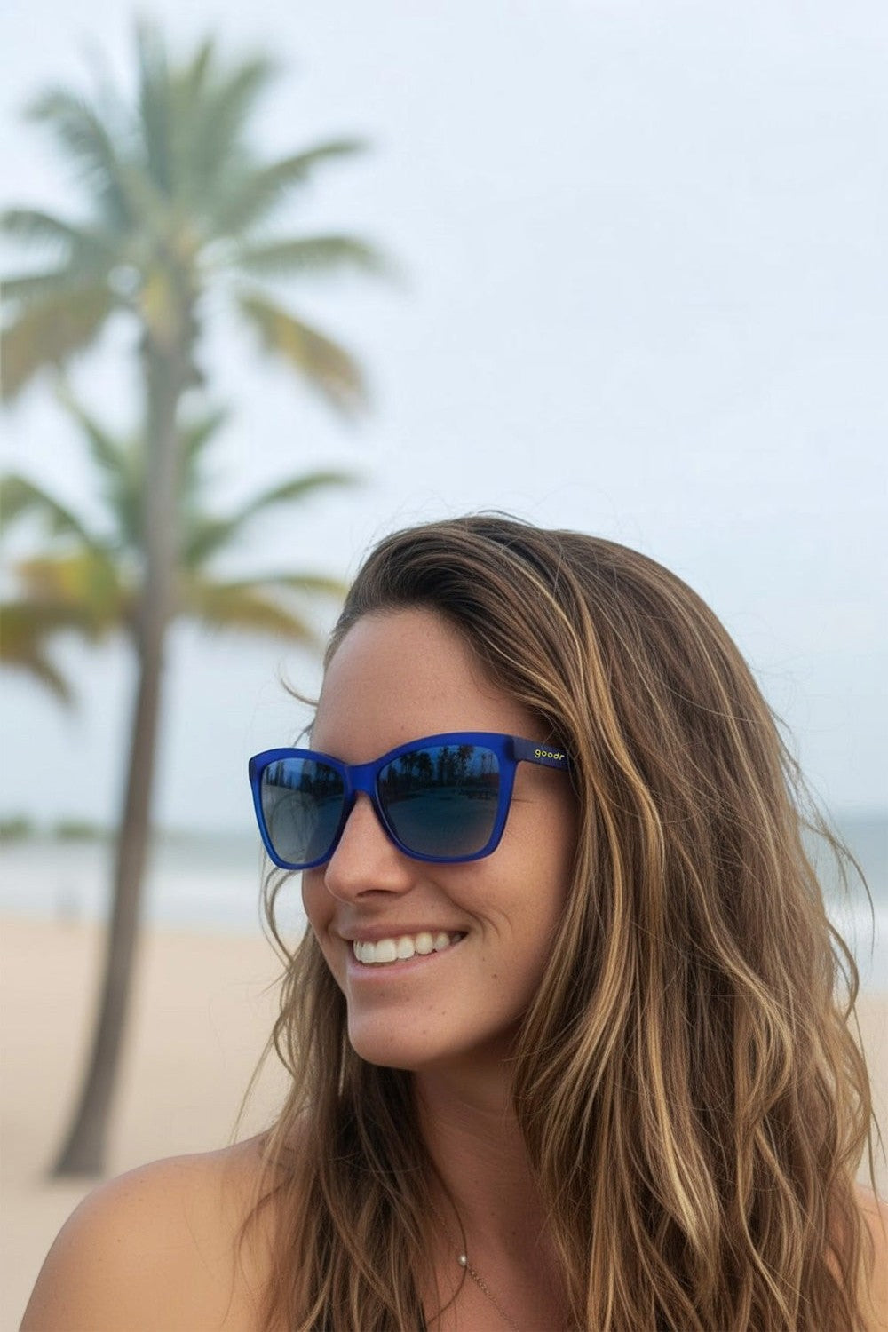 Mulher usando óculos de sol SPORTBR Goodr Pop Art Prodigy azul com lentes polarizadas em cenário de praia.
