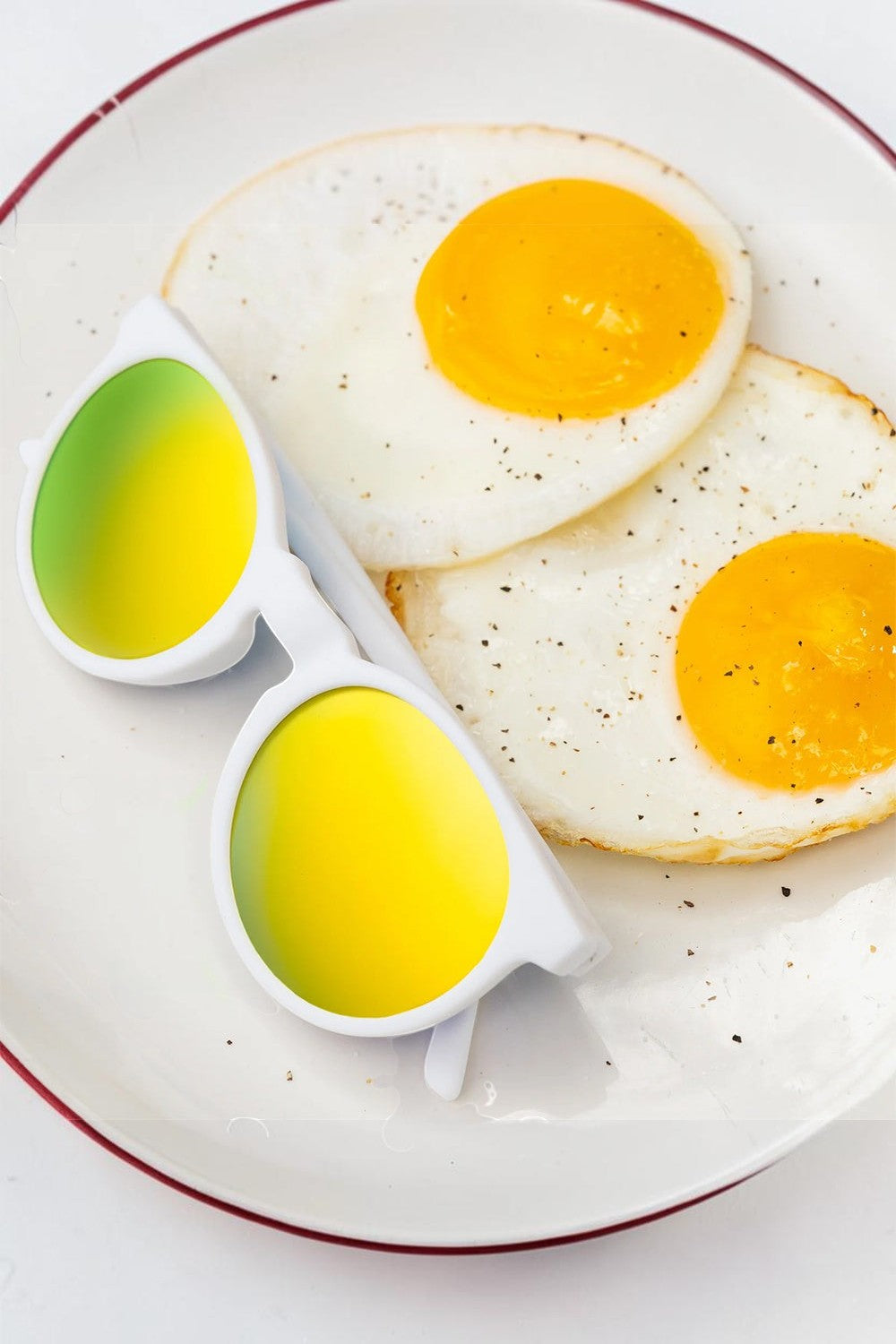Óculos de sol SPORTBR Goodr Yolk's on You com armação branca e lentes espelhadas amarelas sobre um prato com ovos fritos.