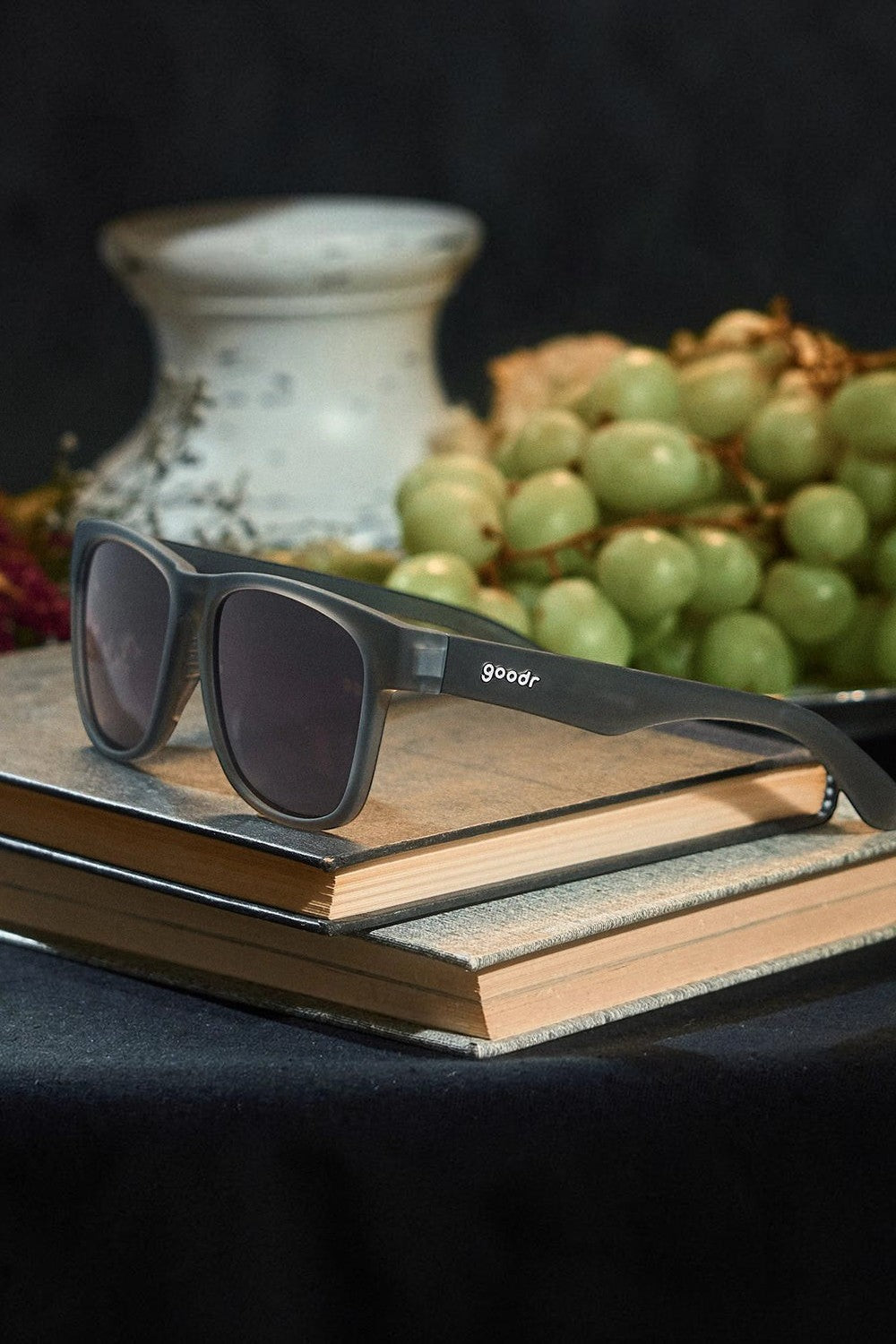 Óculos de sol SPORTBR Goodr Always the Main Event cinza com lentes polarizadas sobre livros e uvas ao fundo
