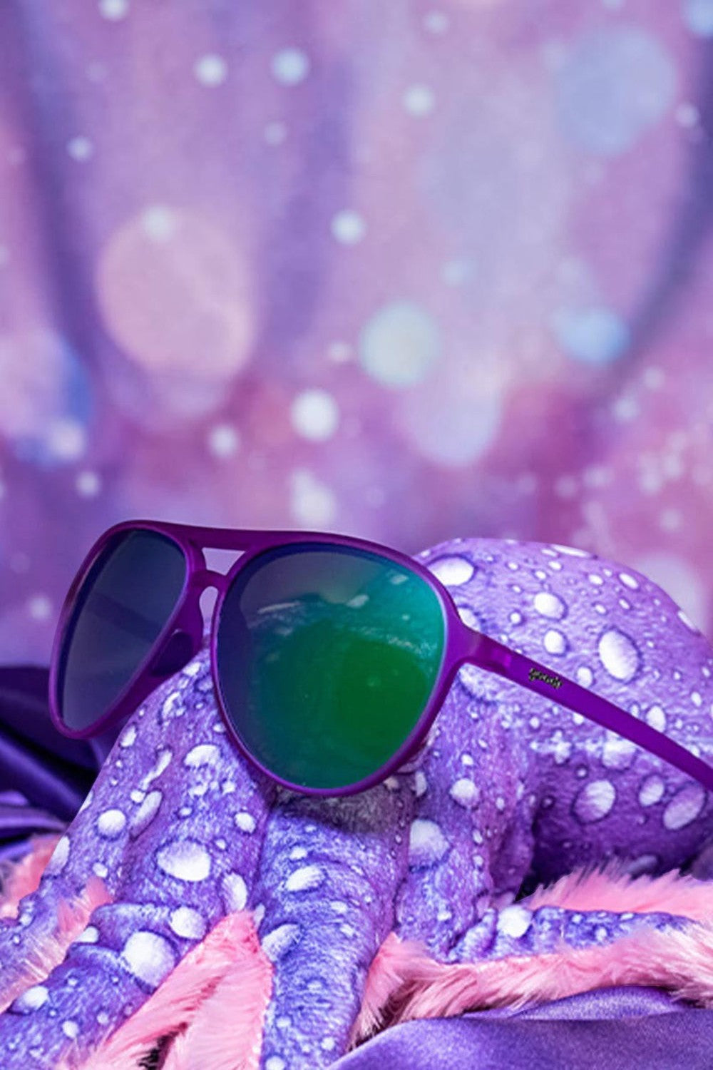 Óculos de sol SPORTBR Goodr lilás com lentes espelhadas verdes, ideal para corrida, sobre um polvo de pelúcia.