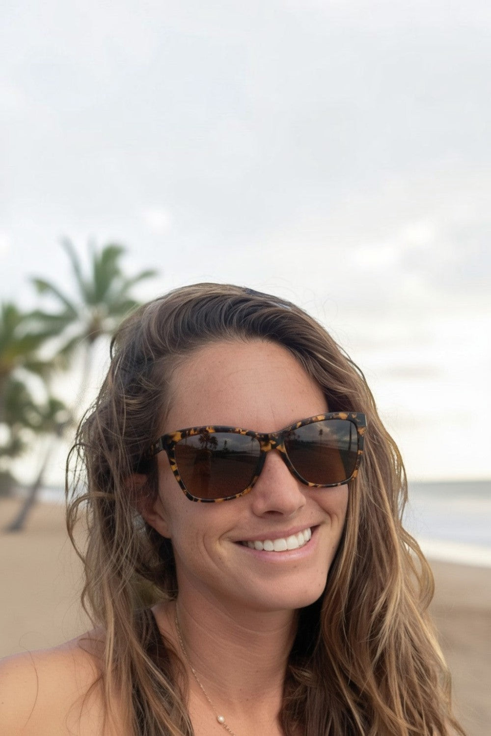 Mulher sorrindo na praia usando óculos de sol SPORTBR Goodr Vanguard Visionary com armação marrom e lentes polarizadas.