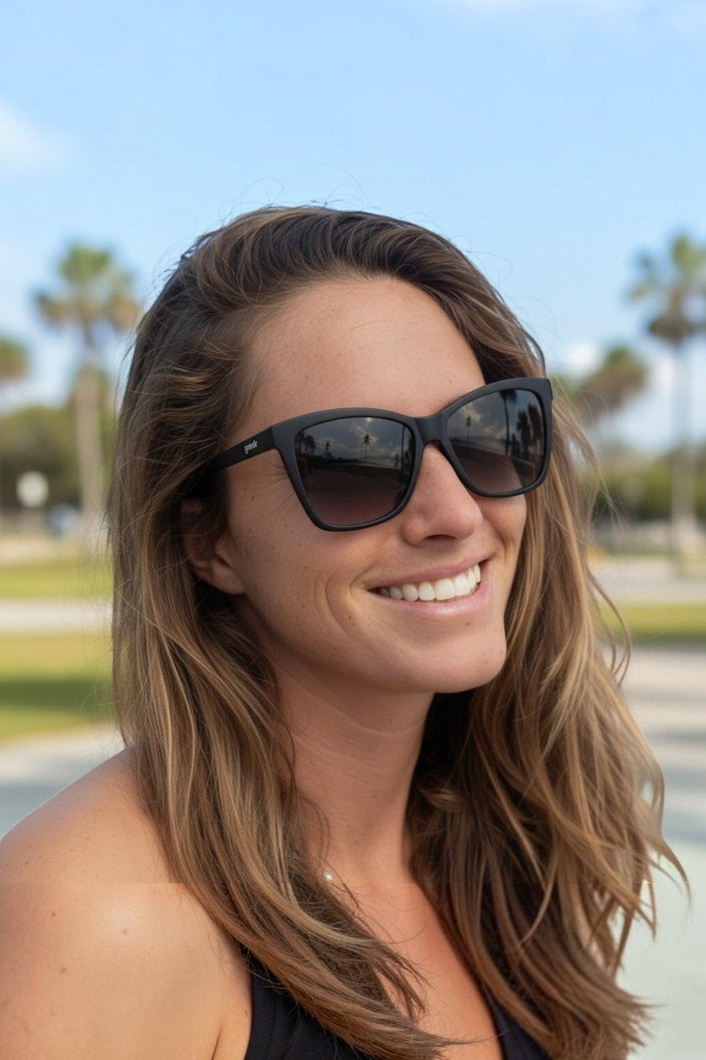 Mulher sorrindo usando óculos de sol SPORTBR Goodr New Wave Renegade pretos com lentes polarizadas em ambiente externo