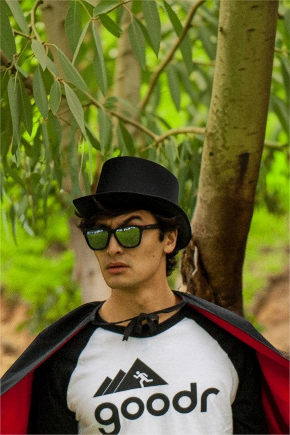 Homem usando óculos de sol SPORTBR Goodr Ron's Smoke and Mirrors, cartola e capa em ambiente de natureza