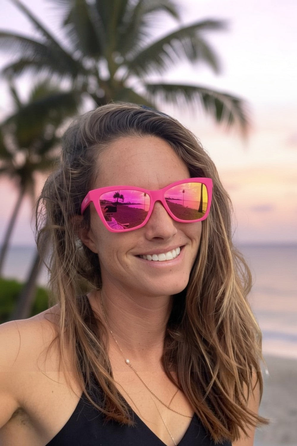 Mulher usando óculos de sol SPORTBR Goodr rosa com lentes espelhadas em praia ao pôr do sol