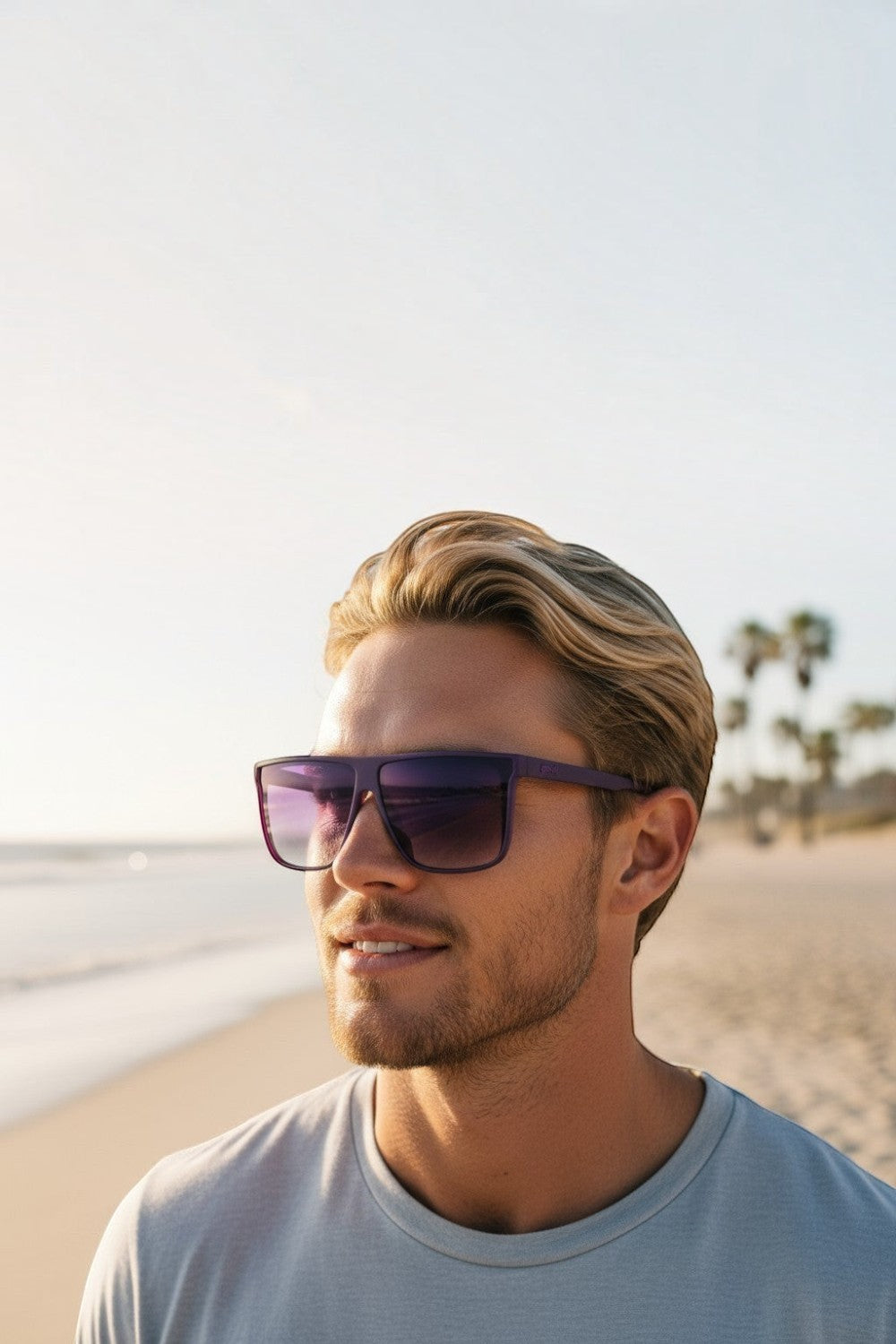 Homem loiro usando óculos de sol SPORTBR Goodr roxos com lentes polarizadas em uma praia ensolarada.