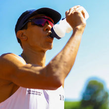 Atleta SPORTBR bebendo água de uma garrafa branca com tampa azul, usando óculos de sol esportivos e boné.