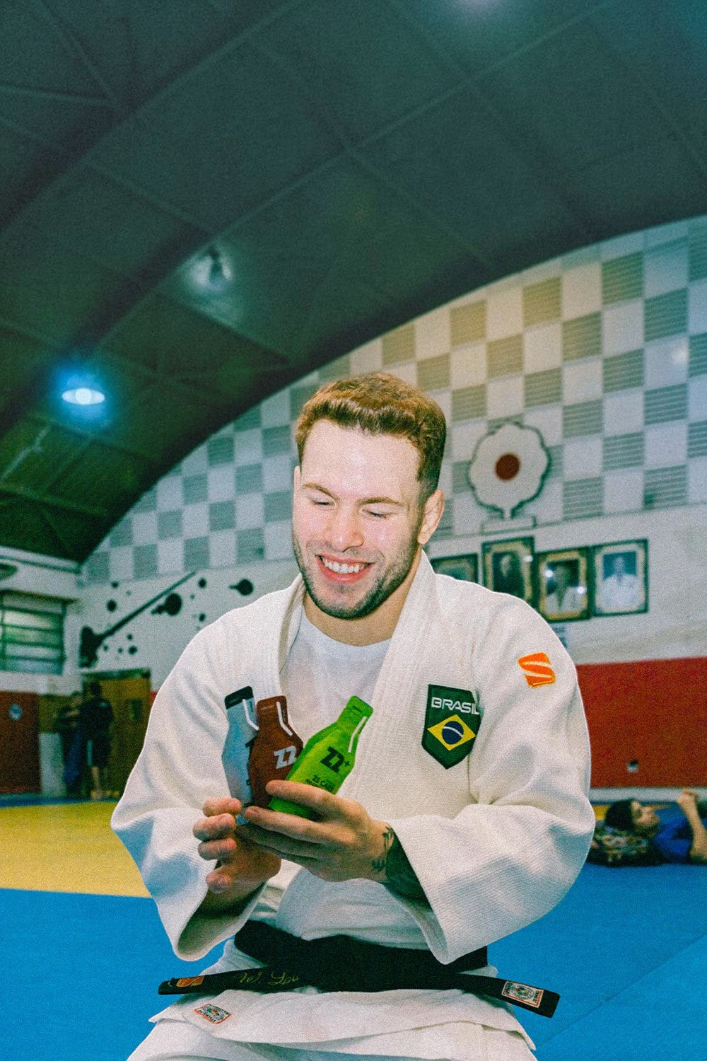 Atleta SPORTBR de judô sorrindo, segurando sachês de gel de carboidrato Z2 em um tatame de academia.