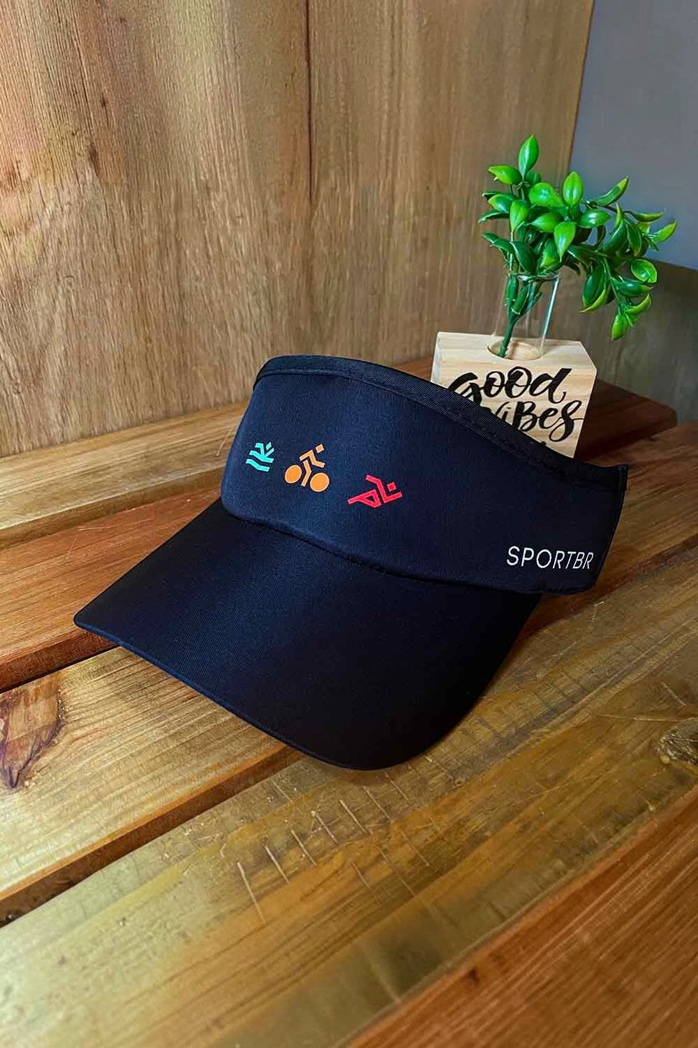 Viseira SPORTBR preta para corrida com ícones coloridos de triatlo e ajuste elástico, sobre superfície de madeira.